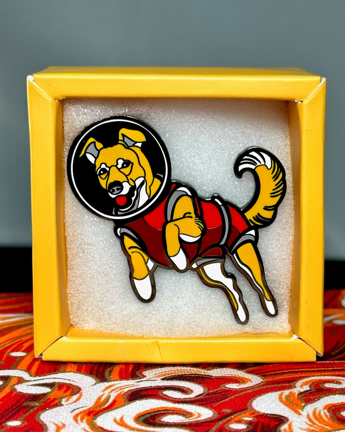 An enamel pin depicting Laika, the dog sent into space in 1957. A light brown/yellow dog, wearing a red space suit and helmet. The pin is in a white foam background inside of a yellow box, sitting on a red, orange and white fabric.