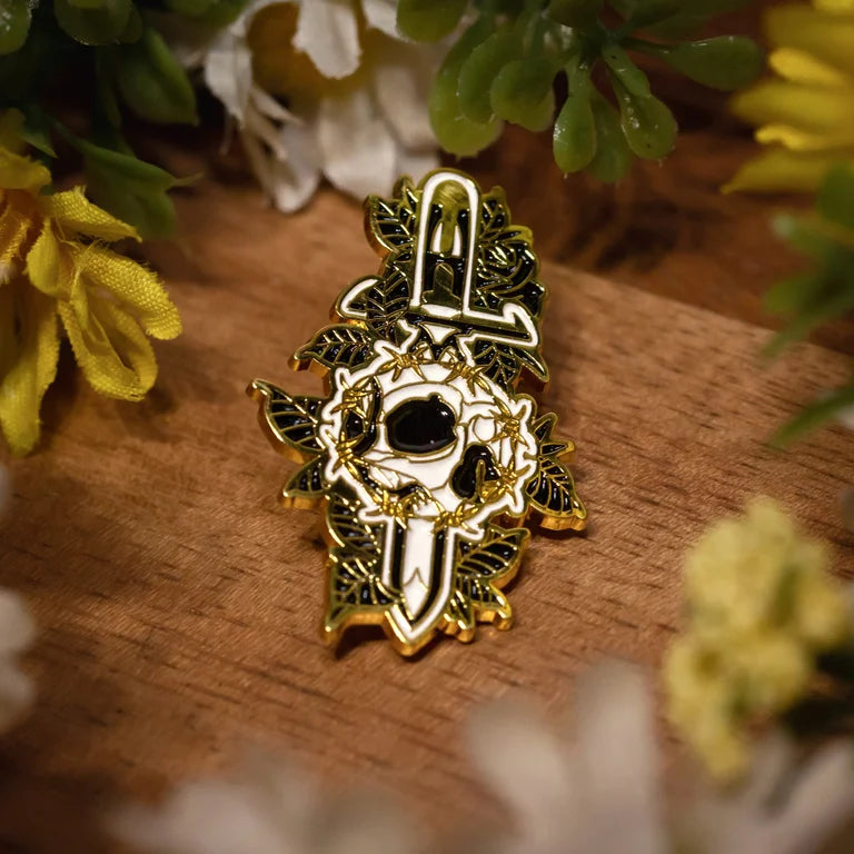 Close up on a gold, black and white dagger with roses behind it. The dagger is piercing a ring of barbed wire with a skull inside of it. The pin is sitting on a wood grain background with small yellow, green and white flower decor