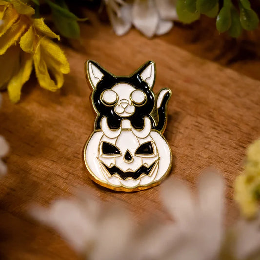 Close up of an enamel pin of a white jack-o-lantern with a black and white tuxedo cat popping out from the top. Wood grain background with small yellow, green and white flower decor