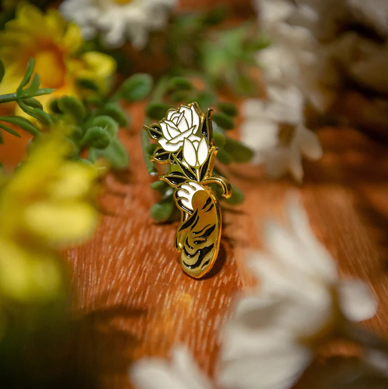 Close up of an enamel pin of a gold cat paw/arm with black stripes holding a white, black and gold flower. Wood grain background with small yellow, green and white flower decor