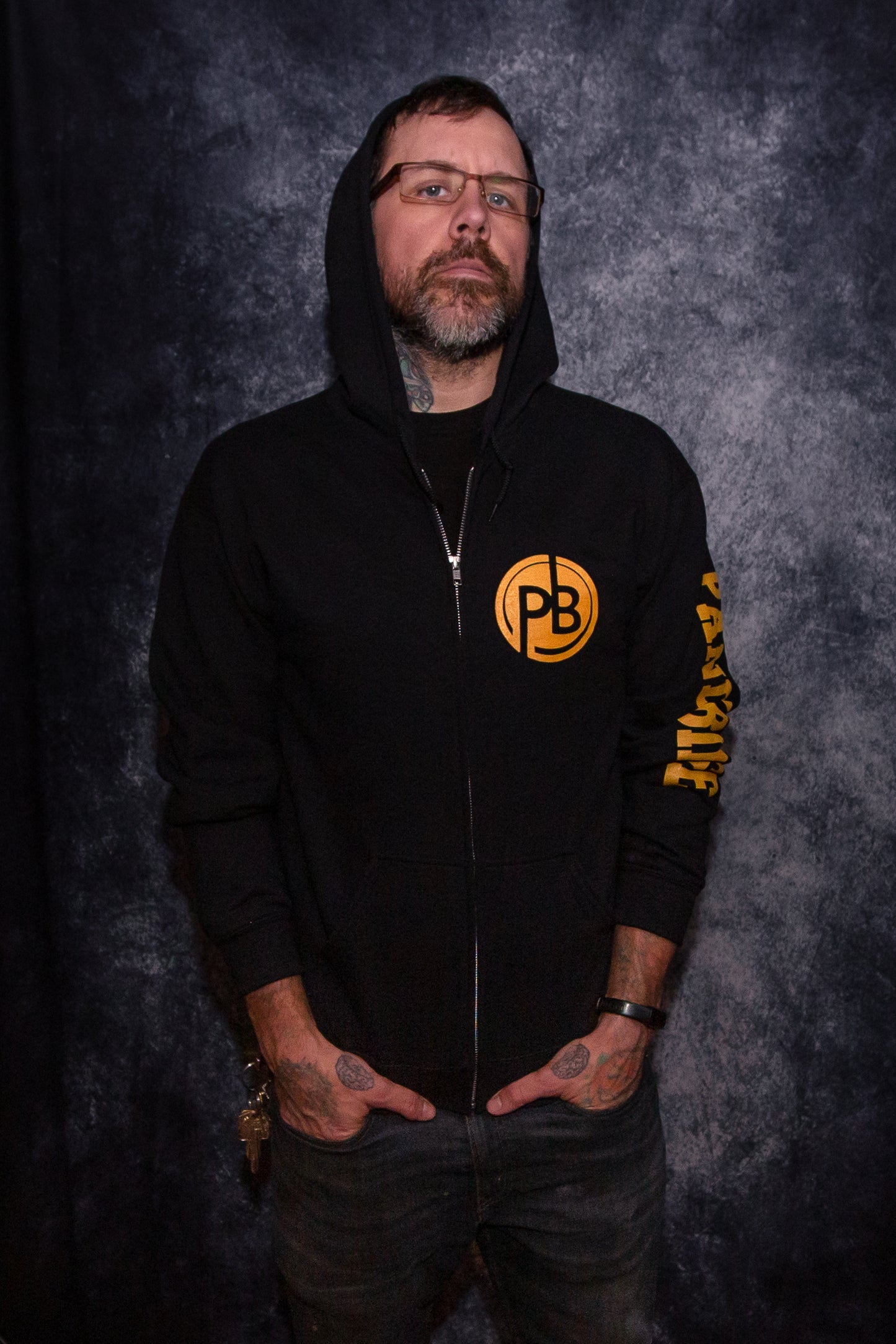 Man wearing a black zip up hoodie in front of a gray photo backdrop. The hoodie features the round  "PB" PandaBinge logo in gold on the left chest. The words "PandaBinge" are printed in gold down the left sleeve in all capital letters and in a block font. 