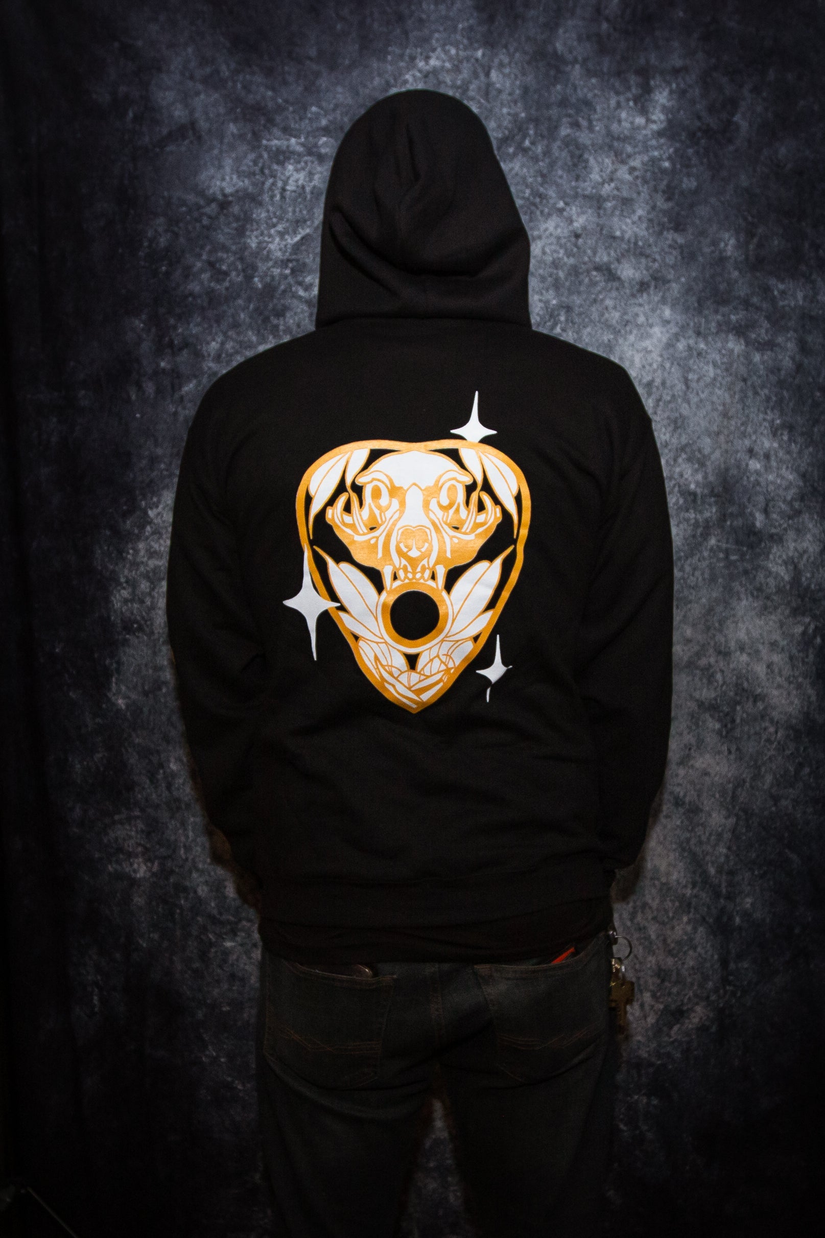 Man wearing a black hoodie in front of a gray photo backdrop, standing backwards to show the design on the back of the sweatshirt. The back of the hoodie features PandaBinge Cat Planchette design. This design is a black-colored planchette with a white cat skull and white leaves, gold outline and three white sparkles/stars.