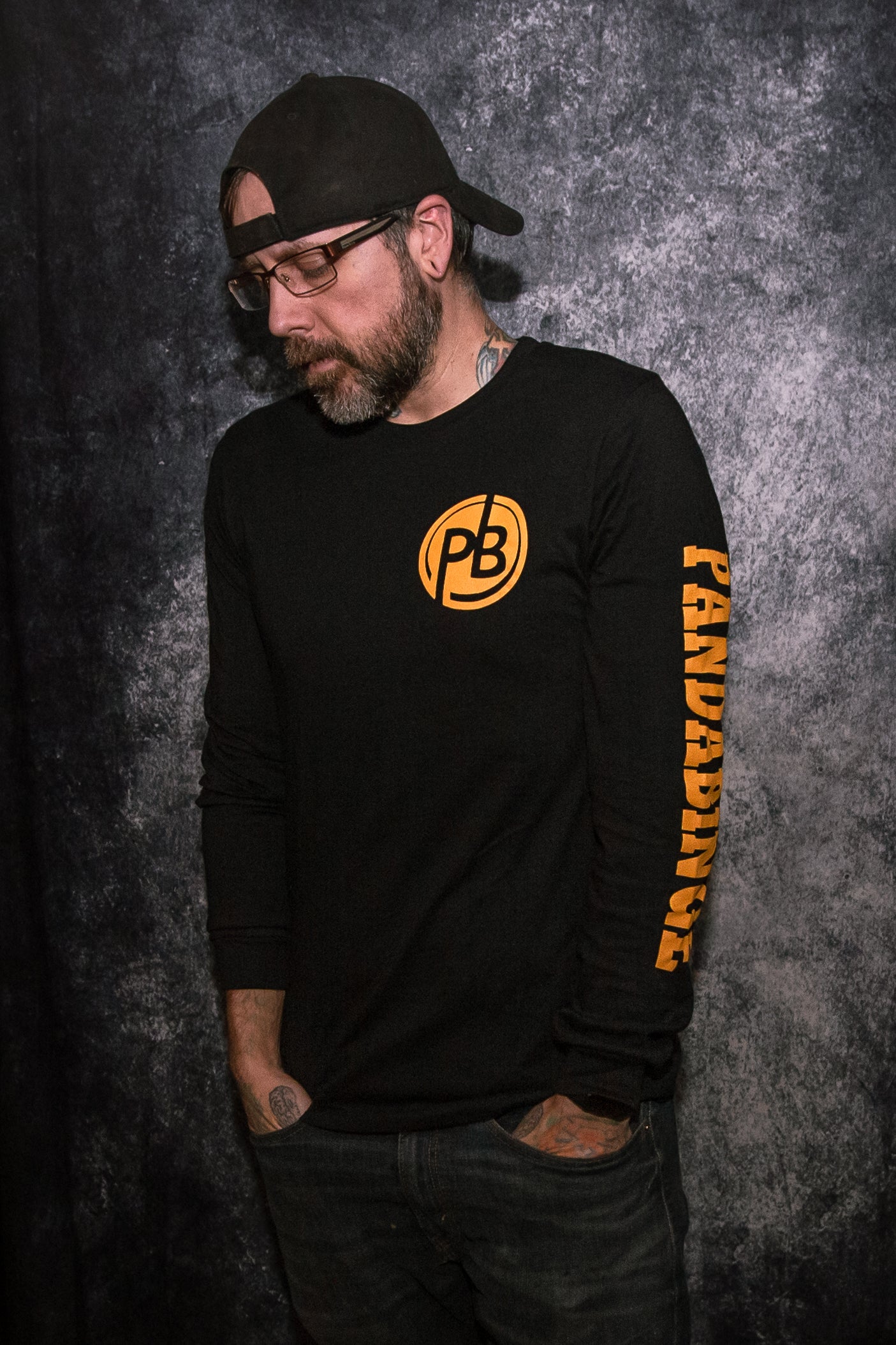 Man wearing a black long sleeve t-shirt in front of a gray photo backdrop. The shirt features the round  "PB" PandaBinge logo in gold on the left chest. The words "PandaBinge" are printed in gold down the left sleeve in all capital letters and in a block font. 