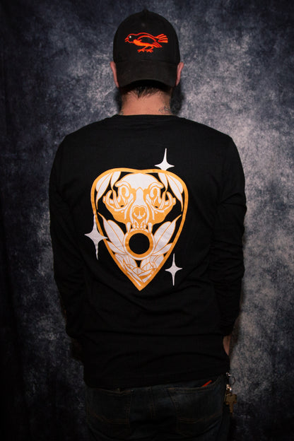 Man wearing a black long sleeve shirt in front of a gray photo backdrop, standing with his back to the camera to show off the back of the shirt. The back of the shirt features PandaBinge Cat Planchette design. This design is a black-colored planchette with a white cat skull and white leaves, gold outline and three white sparkles/stars.