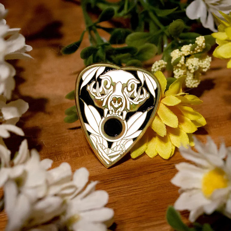 Close up of an enamel pin of a black and white cat skull with leaves on a planchette. Wood grain background with small yellow, green and white flower decor