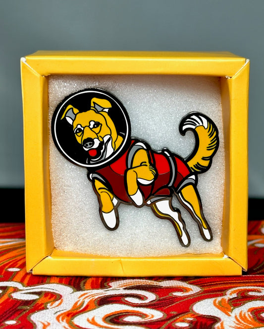 An enamel pin depicting Laika, the dog sent into space in 1957. A light brown/yellow dog, wearing a red space suit and helmet. The pin is in a white foam background inside of a yellow box, sitting on a red, orange and white fabric. 