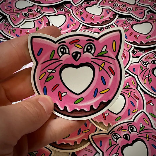 A donut drawing that looks like a cat - pink icing donut sprinkles, cat eyes and nose at the top and ears. Refrigerator magnet in hand, background is a pile of the same magnet.